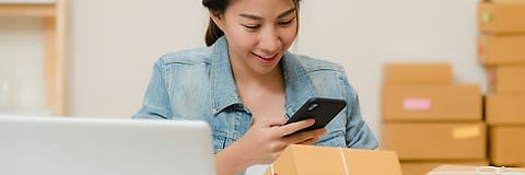 A shop owner checking orders on a phone beside a stack of parcels.